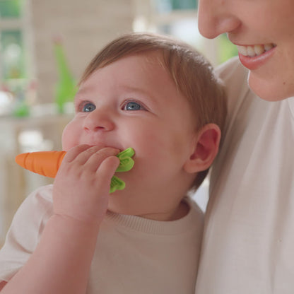 A smiling baby, dressed in white and held by an adult, plays with a Nuby Carrot Teether from the Carrot Teether Set (2 Pack) in a brightly lit room.