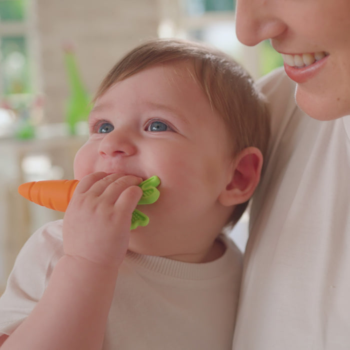 A smiling baby chews on the Nuby Sensory Carrot Teether for relief while being held by an adult in a white shirt. Both look happy in a bright, cozy indoor setting.